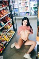 A woman in a pink shirt and black thong sitting on the floor in front of a refrigerator.